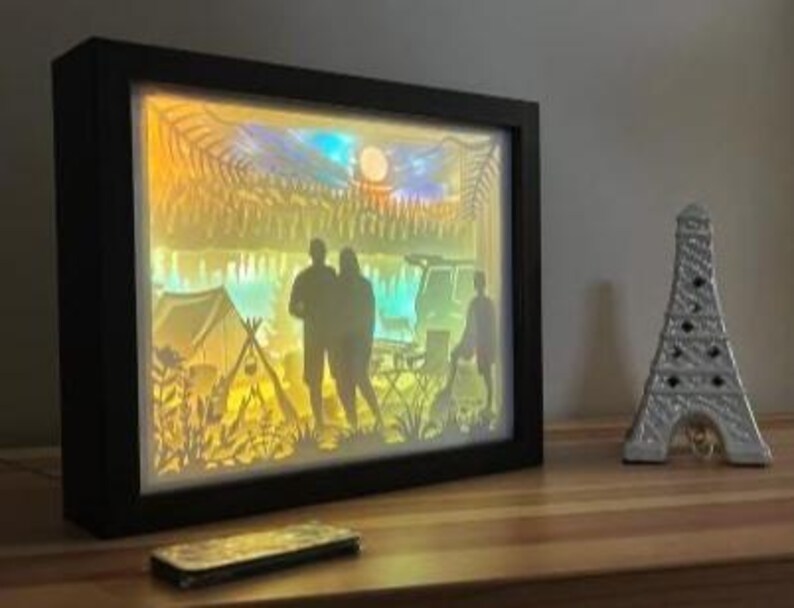 May include: A rectangular black shadow box frame containing a multi-layered paper cutout scene of a camping trip. The backlit display features silhouettes of two people standing near a tent, a vehicle, and trees under a moonlit sky with warm yellow and blue lighting.