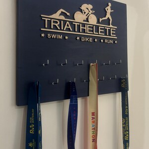K&ouml;nnte beinhalten: Ein dunkelblaues Holzmedaillen-Display mit den Worten "TRIATHELETE", "SWIM", "BIKE" und "RUN" in goldenen Buchstaben. Das Display hat Haken zum Aufh&auml;ngen von Medaillen. Drei Medaillen h&auml;ngen an den Haken, eine blaue, eine gelbe und eine rote.