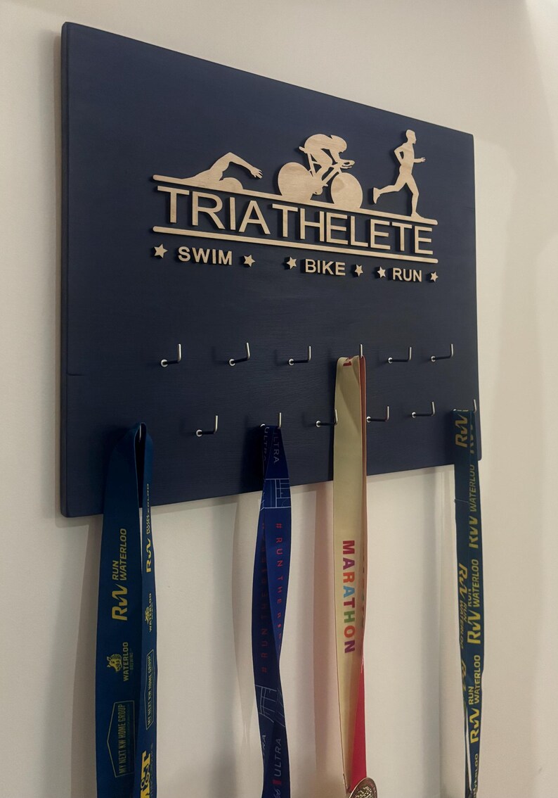 K&ouml;nnte beinhalten: Ein dunkelblaues Holzmedaillen-Display mit den Worten "TRIATHELETE", "SWIM", "BIKE" und "RUN" in goldenen Buchstaben. Das Display hat Haken zum Aufh&auml;ngen von Medaillen. Drei Medaillen h&auml;ngen an den Haken, eine blaue, eine gelbe und eine rote.
