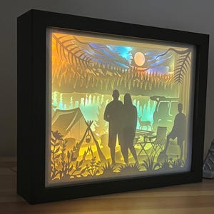 May include: A rectangular shadow box light featuring a layered paper cutout scene of a camping site at night. The backlit display shows silhouettes of two adults and a child, a tent, a campfire, a deer, and pine trees under a moonlit sky.