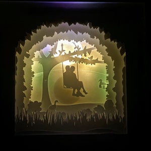 K&ouml;nnte beinhalten: Eine wei&szlig;e Silhouette eines Paares, das auf einer Schaukel unter einem Baum mit einem Vollmond im Hintergrund sitzt. Die Szene wird von hinten beleuchtet und erzeugt einen sanften Schein.