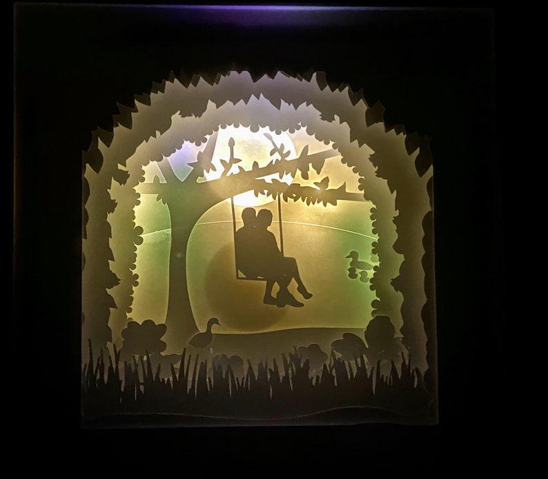 K&ouml;nnte beinhalten: Eine wei&szlig;e Silhouette eines Paares, das auf einer Schaukel unter einem Baum mit einem Vollmond im Hintergrund sitzt. Die Szene wird von hinten beleuchtet und erzeugt einen sanften Schein.