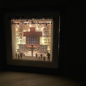 May include: A light-up shadow box with a hospital scene, featuring a silhouette of a nurse with wings, a hospital building, and a family. The text "Not All Angels Have Wings Some Have Stethoscopes" is displayed above the scene.