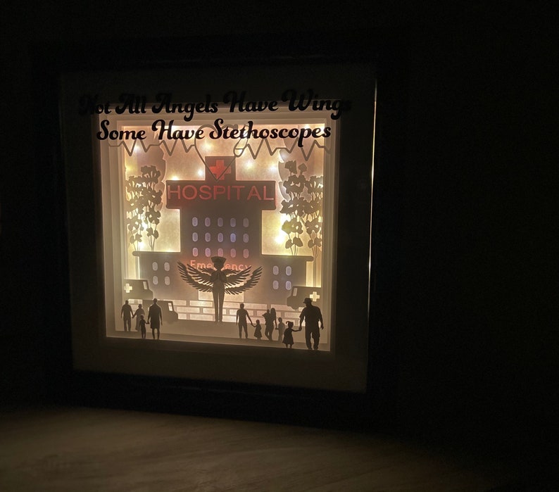 May include: A light-up shadow box with a hospital scene, featuring a silhouette of a nurse with wings, a hospital building, and a family. The text "Not All Angels Have Wings Some Have Stethoscopes" is displayed above the scene.