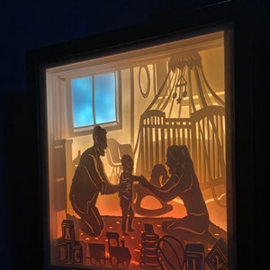 Op de afbeelding: Een schaduwdoos van papier met een gezinsscene. De silhouetten van een vader, moeder en kind spelen in een kinderkamer met een wieg, speelgoed en een schommelpaard. De sc&egrave;ne wordt van achteren verlicht, waardoor een warme gloed ontstaat.