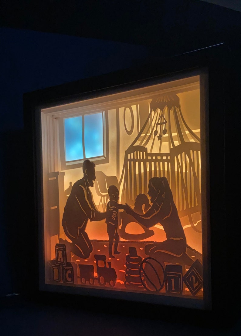 Op de afbeelding: Een schaduwdoos van papier met een gezinsscene. De silhouetten van een vader, moeder en kind spelen in een kinderkamer met een wieg, speelgoed en een schommelpaard. De sc&egrave;ne wordt van achteren verlicht, waardoor een warme gloed ontstaat.