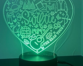Może przedstawiać: Nocna lampka LED w kształcie serca z wygrawerowanym słowem "Swiftie" i "1989" w akrylu. Projekt zawiera gitarę, motyle, kapelusz kowbojski i inne ikony. Światło jest zielone i stoi na czarnej podstawie.