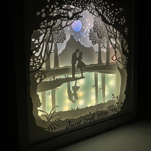 K&ouml;nnte beinhalten: Ein wei&szlig;es Papierkunstwerk mit einem Paar, das in einer Waldszene silhouettiert ist. Die Szene umfasst einen See, B&auml;ume, einen Berg, einen Mond und Sterne. Das Kunstwerk ist mit LED-Lichtern beleuchtet und tr&auml;gt den Text "June 2nd, 2018" am unteren Rand.
