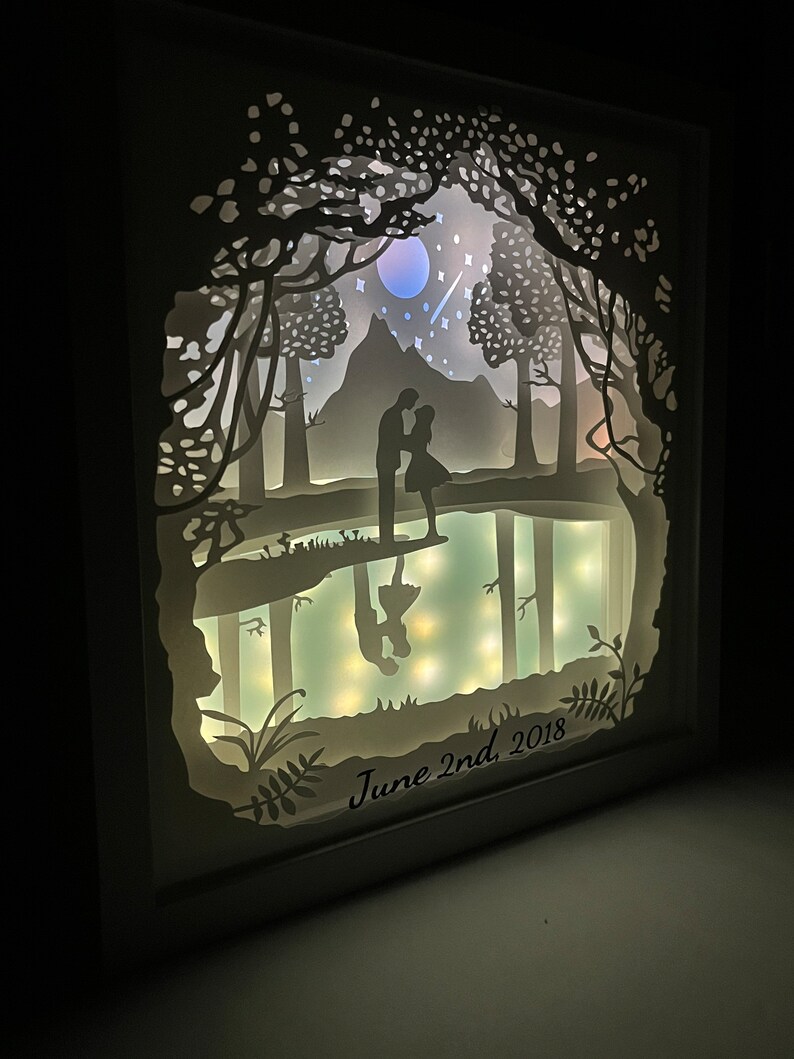 K&ouml;nnte beinhalten: Ein wei&szlig;es Papierkunstwerk mit einem Paar, das in einer Waldszene silhouettiert ist. Die Szene umfasst einen See, B&auml;ume, einen Berg, einen Mond und Sterne. Das Kunstwerk ist mit LED-Lichtern beleuchtet und tr&auml;gt den Text "June 2nd, 2018" am unteren Rand.