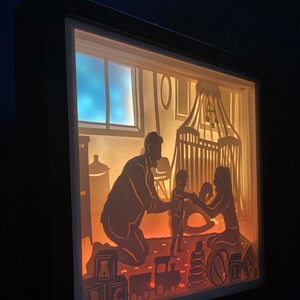Op de afbeelding: Een schaduwdoos van papier met een gezinsscene. Een man en een vrouw knielen op een tapijt met een kind dat tussen hen staat. Het kind houdt een speelgoed vast. De sc&egrave;ne wordt van achteren verlicht, waardoor een warme gloed ontstaat. Er liggen speelgoedjes op de vloer, waaronder blokken die "ABC" spellen en een speelgoedtrein.