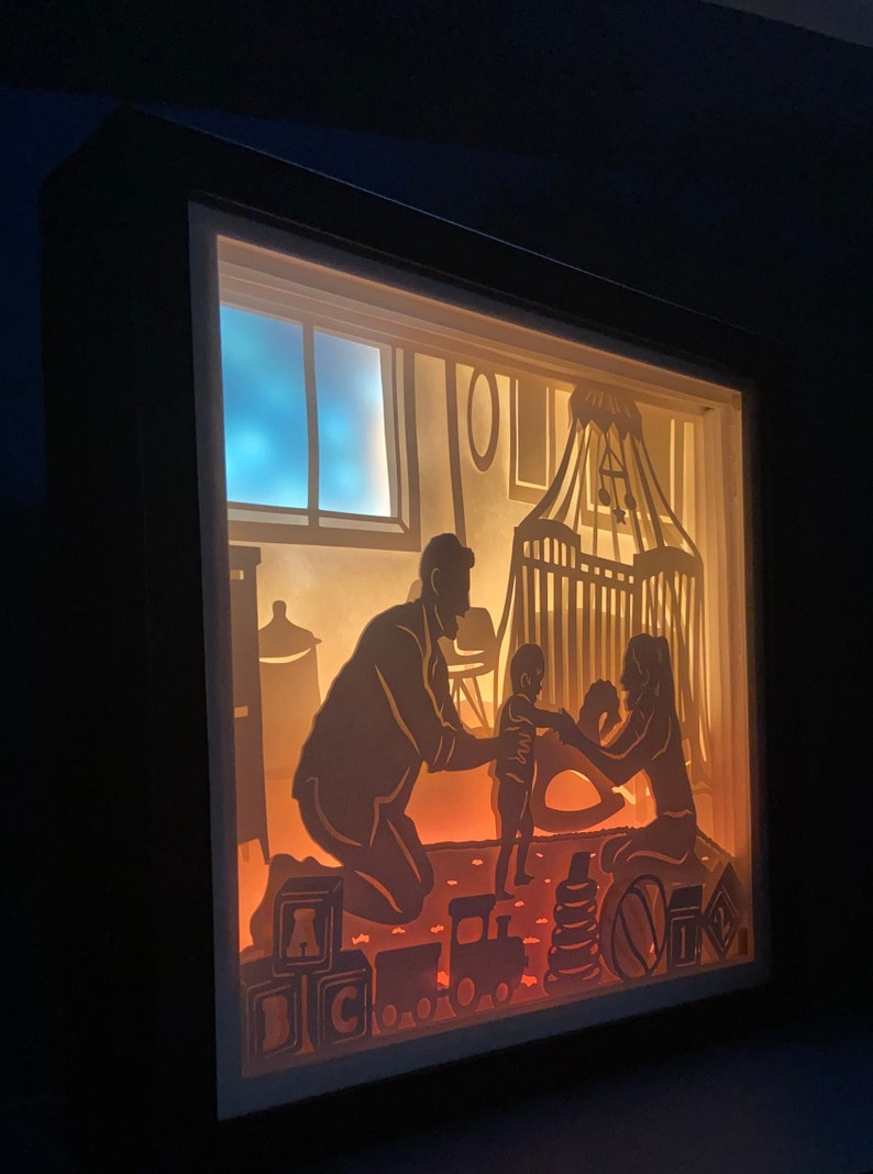 Op de afbeelding: Een schaduwdoos van papier met een gezinsscene. Een man en een vrouw knielen op een tapijt met een kind dat tussen hen staat. Het kind houdt een speelgoed vast. De sc&egrave;ne wordt van achteren verlicht, waardoor een warme gloed ontstaat. Er liggen speelgoedjes op de vloer, waaronder blokken die "ABC" spellen en een speelgoedtrein.