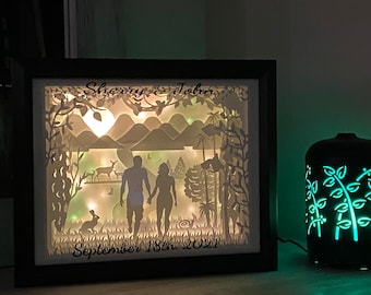 Könnte beinhalten: Ein weißes Papier-Schnittbild in einem schwarzen Rahmen mit einer beleuchteten Szene eines Paares, das durch einen Wald mit Hirschen, einem Kaninchen und Bergen im Hintergrund geht. Der Text "Sherry & John September 18th 2021" befindet sich am unteren Rand des Designs.