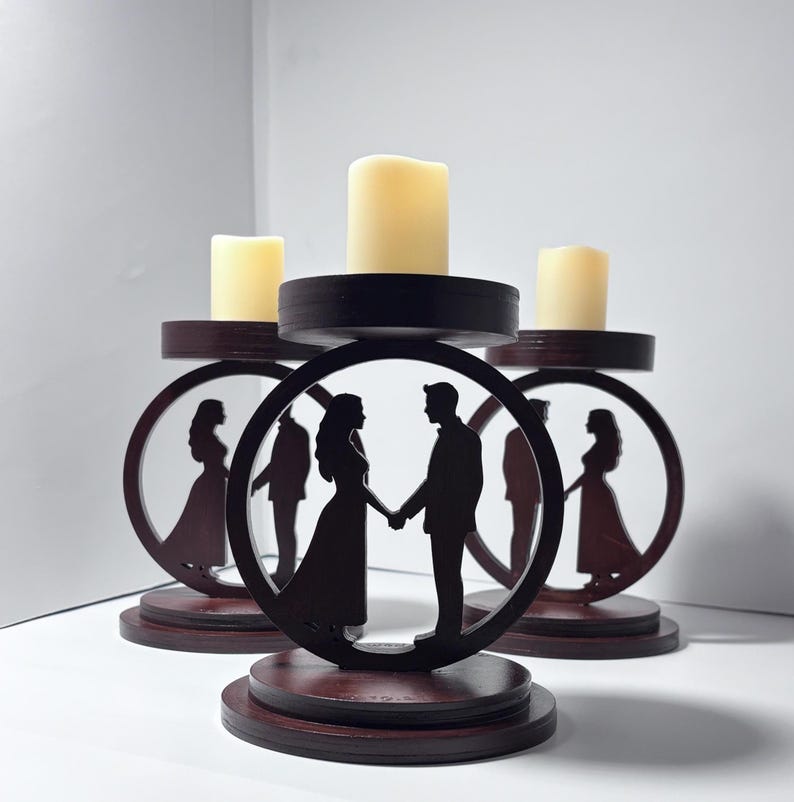 K&ouml;nnte beinhalten: Drei h&ouml;lzerne Kerzenhalter mit einem kreisf&ouml;rmigen Design, das eine Silhouette eines Paares zeigt, das sich an den H&auml;nden h&auml;lt. Jeder Halter hat einen dunkelbraunen Sockel und eine kreisf&ouml;rmige Oberseite mit einer cremefarbenen Kerze. Ideal f&uuml;r Hochzeiten.