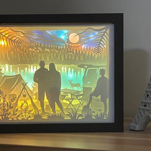 May include: A rectangular shadow box with a black frame containing a multi-layered paper cut scene of a camping site at night. The illuminated display features silhouettes of two people, a tent, a campfire, a deer, and a vehicle under a moonlit sky.