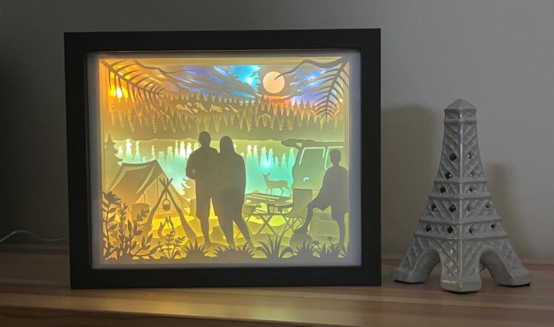 May include: A rectangular shadow box with a black frame containing a multi-layered paper cut scene of a camping site at night. The illuminated display features silhouettes of two people, a tent, a campfire, a deer, and a vehicle under a moonlit sky.