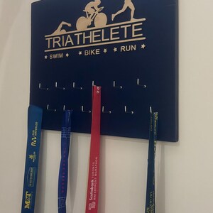 K&ouml;nnte beinhalten: Ein dunkelblaues, h&ouml;lzernes Medaillen-Pr&auml;sentationsbrett mit den Worten "TRIATHELETE" und "* SWIM * BIKE * RUN *" in goldenen Lettern. Das Brett hat Haken zum Aufh&auml;ngen von Medaillen. Drei Medaillen h&auml;ngen an den Haken, eine blaue, eine rote und eine goldene.