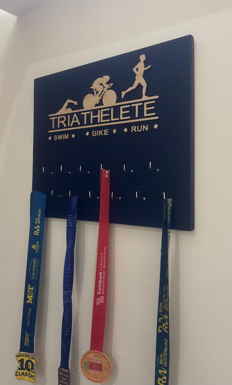 K&ouml;nnte beinhalten: Ein dunkelblaues, h&ouml;lzernes Medaillen-Pr&auml;sentationsbrett mit den Worten "TRIATHELETE" und "* SWIM * BIKE * RUN *" in goldenen Lettern. Das Brett hat Haken zum Aufh&auml;ngen von Medaillen. Drei Medaillen h&auml;ngen an den Haken, eine blaue, eine rote und eine goldene.