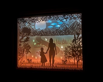 Puede incluir: Una obra de arte de papel recortado que representa a una madre y un hijo caminando por un paisaje de bosque. La escena está iluminada desde atrás, creando un brillo cálido. El bosque está lleno de árboles, un camino sinuoso y una luna llena en el cielo.