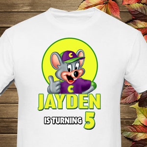 Puede incluir: Camiseta blanca con un ratón de dibujos animados que lleva una camisa morada y un sombrero verde. El ratón está haciendo un pulgar hacia arriba. El texto "JAYDEN IS TURNING 5" está impreso en amarillo y verde en la camiseta.
