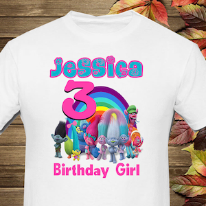 May include: White t-shirt with a pink "3" and a rainbow behind it. The shirt also has the name "Jessica" in blue with pink flowers and the words "Birthday Girl" in pink. The shirt features a group of Trolls characters.