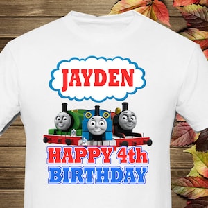 May include: White t-shirt featuring a blue, red and white cloud with the name "JAYDEN" in red lettering. Below the cloud are three cartoon trains, Thomas, Percy and James, with the text "HAPPY 4th BIRTHDAY" in red and blue lettering.