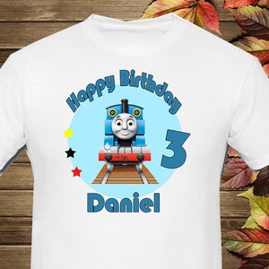Puede incluir: Camiseta blanca con un círculo azul que presenta a Thomas la locomotora, las palabras "Feliz cumpleaños", el número "3" y el nombre "Daniel".