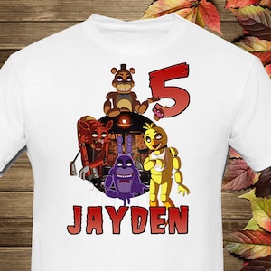 以下が含まれることがあります： 白いTシャツ。アニメのキャラクター、数字の5、そして「JAYDEN」という名前が赤色で描かれたカラフルなグラフィック。茶色のクマ、赤いキツネ、紫のウサギ、黄色のニワトリが含まれています。