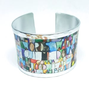 Puede incluir: Un brazalete de metal plateado con un diseño impreso colorido que incluye palabras como "words", "cute", "down", "gonna", "good", "team", "brave", y más.