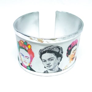 May include: Silver-toned cuff bracelet featuring three portraits of Frida Kahlo. The portraits are in vibrant colors and black and white, set against a white background. The bracelet has a hammered texture on the inside.