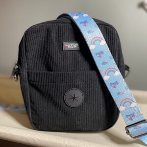 Puede incluir: Bolso bandolera de pana negro con una correa azul claro con arco&iacute;ris, patines y estrellas. El bolso tiene un bolsillo frontal y un emblema circular negro. La etiqueta dice "Luna & Lady Design Co."