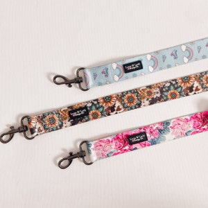 Puede incluir: Tres correas estampadas con cierres met&aacute;licos negros. Una es azul claro con arco&iacute;ris y patines. Otra tiene un dise&ntilde;o floral y de piel de vaca. La tercera correa es rosa con estampados florales. Texto "Luna & Lady Design Co."