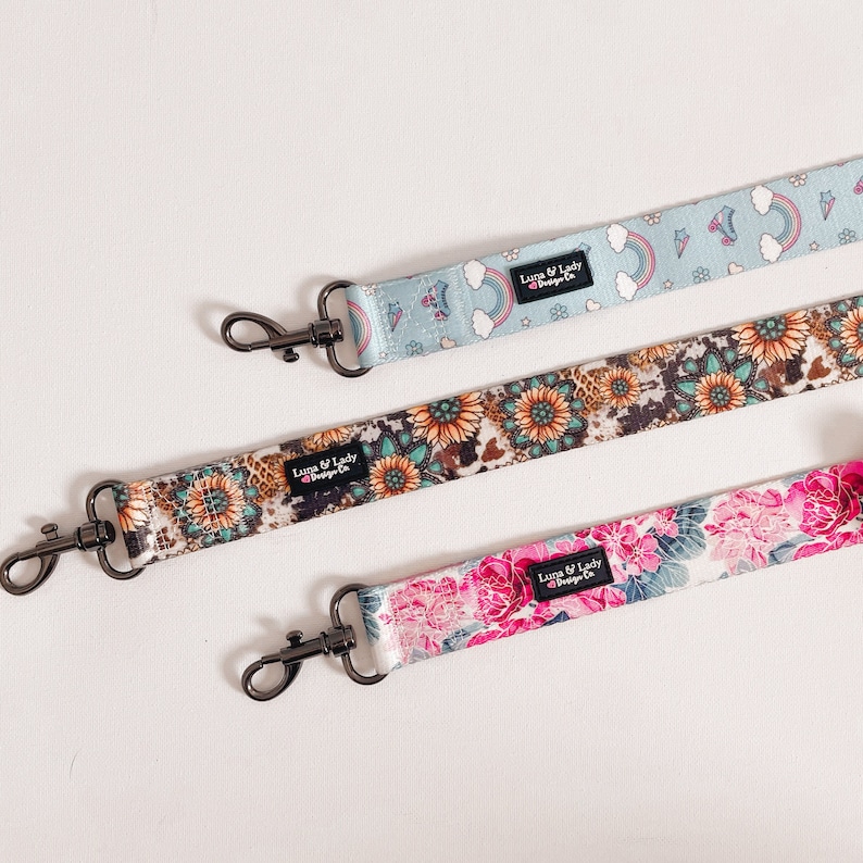 Puede incluir: Tres correas estampadas con cierres met&aacute;licos negros. Una es azul claro con arco&iacute;ris y patines. Otra tiene un dise&ntilde;o floral y de piel de vaca. La tercera correa es rosa con estampados florales. Texto "Luna & Lady Design Co."