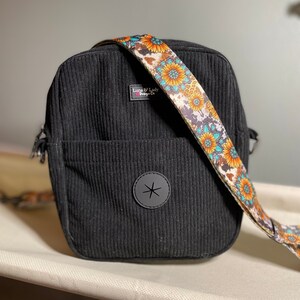 Puede incluir: Bolso bandolera de pana negro con una correa estampada con girasoles. El bolso tiene un bolsillo frontal y un parche circular negro con un dise&ntilde;o de estrella. La correa presenta girasoles, detalles turquesa y un estampado de piel de vaca.