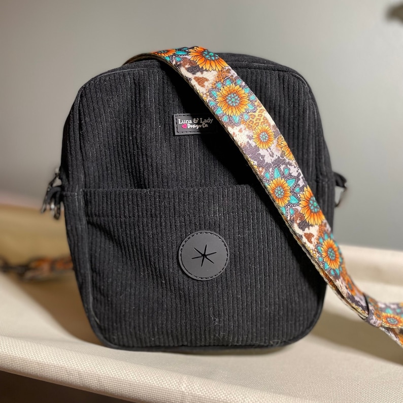 Puede incluir: Bolso bandolera de pana negro con una correa estampada con girasoles. El bolso tiene un bolsillo frontal y un parche circular negro con un dise&ntilde;o de estrella. La correa presenta girasoles, detalles turquesa y un estampado de piel de vaca.