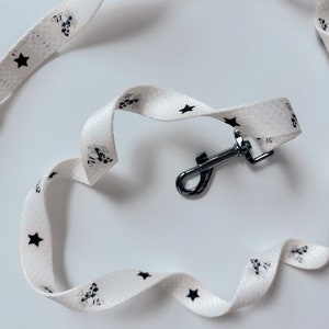 Könnte beinhalten: Weiße Hundeleine mit einem Muster aus schwarzen Sternen und Pfotenabdrücken. Sie hat einen silberfarbenen Metallverschluss. Die Leine besteht aus einem weichen, flexiblen Material.