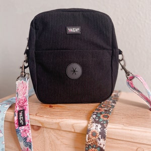 Puede incluir: Bolso de pana negro con un bolsillo frontal y un emblema de estrella gris. El bolso tiene forma rectangular con cierre de cremallera superior y una correa desmontable con dise&ntilde;os florales y estampados. El bolso tiene el texto "Luna & Lady Design Co.".