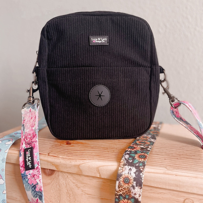 Puede incluir: Bolso de pana negro con un bolsillo frontal y un emblema de estrella gris. El bolso tiene forma rectangular con cierre de cremallera superior y una correa desmontable con dise&ntilde;os florales y estampados. El bolso tiene el texto "Luna & Lady Design Co.".