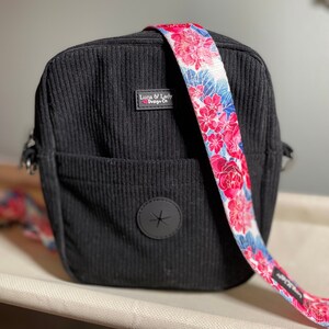 Puede incluir: Bolso bandolera de pana negro con una correa floral. La correa presenta un fondo blanco con flores rosas y rojas y detalles azules. El bolso tiene un bolsillo frontal y un emblema circular negro.