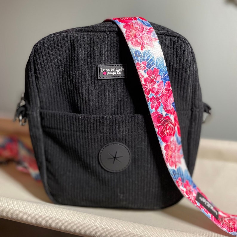 Puede incluir: Bolso bandolera de pana negro con una correa floral. La correa presenta un fondo blanco con flores rosas y rojas y detalles azules. El bolso tiene un bolsillo frontal y un emblema circular negro.