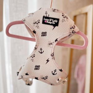 May include: A white dog harness with a black and white checkered pattern. The harness features a black anchor, lightning bolt, and Mickey Mouse print. The text "Good Vibes" and "Luna & Lady Design Co." are printed on the harness.