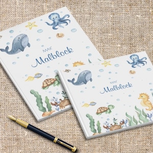 Op de afbeelding: Twee witte notitieboekjes met aquarel illustraties van zeeleven, waaronder walvissen, octopussen en zeepaardjes. De notitieboekjes dragen de naam "MAX Malblock" in blauw schrift. Een pen met een goud-zwarte schacht staat op de voorgrond.