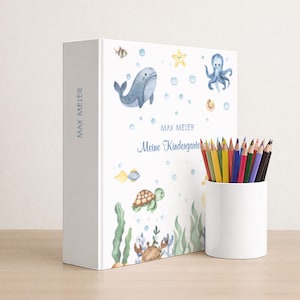 May include: A white book with the text "MAX MEIER" and "Meine Kindergarten" in blue, featuring watercolor illustrations of sea creatures. A white cylindrical pencil holder contains a variety of colored pencils.