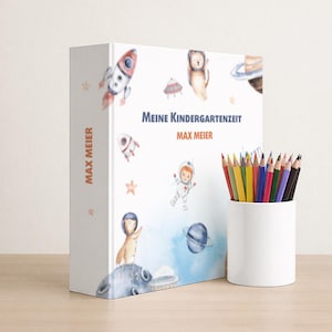 Könnte beinhalten: Ein Kinderbuch mit dem Titel "Meine Kindergartenzeit" von Max Meier, mit Illustrationen von Raketen, Planeten und Astronauten. Ein weißer zylindrischer Behälter enthält eine Sammlung von Farbstiften, die der Szene Farbe verleihen.