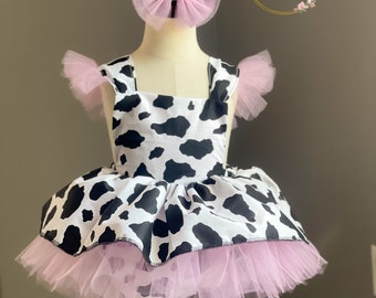 May include: A sleeveless dress featuring a black and white cow print pattern, accented with light pink tulle ruffles on the shoulders and a layered tulle skirt. A matching light pink tulle hair bow is positioned above the dress on the mannequin.