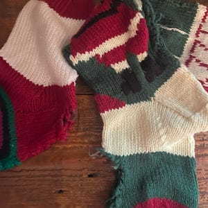 May include: Two knitted Christmas stockings in red, white, and green. One stocking is primarily white with red accents, while the other features a pattern of red, white, and green stripes. The stockings are on a wooden surface.