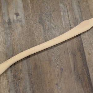 May include: A plain, unfinished wooden axe handle. The handle is light brown and has a slight curve.