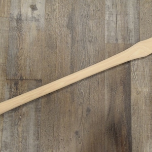 May include: A plain wooden axe handle, approximately 36 inches long, with a slightly flared end. The handle is made of light-colored wood and is lying on a woodgrain surface.