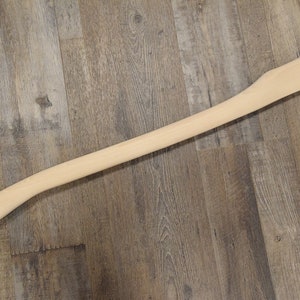 May include: A plain, unfinished wooden axe handle. The handle is light brown and has a smooth surface.