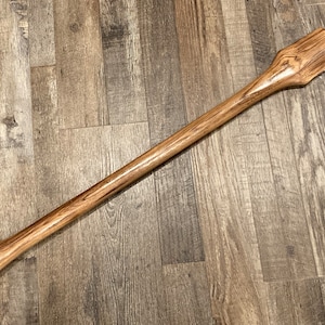 May include: A long, wooden handle for an axe or maul. The handle is made of light brown wood with dark brown streaks. The handle is smooth and has a slightly rounded shape.