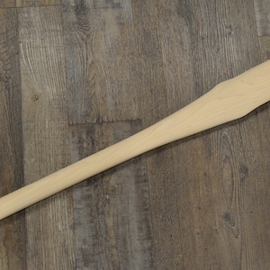 May include: A new, unfinished wooden axe handle, lying diagonally on a wood-grain surface. The handle is a light, natural wood color, with a long, slender shaft and a wider, curved grip area.